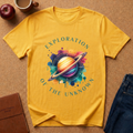 Yellow t-shirt with space-themed design and text Says Exploration of the  unknown on a brown surface with an apple, coffee cup, and jeans. 