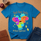 A bright blue graphic t-shirt laid flat on a cork surface, featuring colorful elephant illustrations, the text 'Rock Your Socks' and '# Down Syndrome', plus a blue awareness ribbon. Surrounding props include a coffee mug, apple, notebook, and jeans.