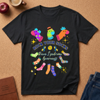 A black short-sleeve graphic T-shirt laid flat on a cork surface, featuring colorful socks and the text 'ROCK YOUR SOCKS' and 'Down Syndrome Awareness.' Surrounding props include a coffee cup, apple, journal, and jeans.