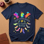 A navy blue graphic t-shirt laid flat on a cork board background, surrounded by lifestyle props including a brown leather journal, a red apple, a coffee cup, and blue jeans. The t-shirt features a colorful graphic of various patterned socks and the text 'ROCK YOUR SOCKS Down Syndrome Awareness'.