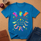 A bright blue graphic T-shirt laid flat on a cork surface, surrounded by a coffee mug, apple, notebook, and jeans. The shirt features colorful sock illustrations and the text 'ROCK YOUR SOCKS Down Syndrome Awareness'.