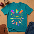 A teal short-sleeve graphic T-shirt laid flat on a cork surface, surrounded by a coffee cup, apple, notebook, pen, and jeans. The shirt features colorful illustrated socks and the text 'ROCK YOUR SOCKS Down Syndrome Awareness'.