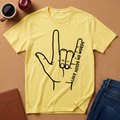 A yellow short-sleeve graphic T-shirt laid flat on a cork surface, featuring a black hand outline with the words 'LOVE NEEDS NO WORDS.' Surrounding props include a coffee mug, an apple, a brown notebook, and blue jeans.