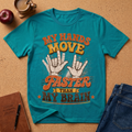 A teal short-sleeve graphic T-shirt laid flat on a cork-like surface, surrounded by props including a coffee cup, apple, notebook, pen, and jeans. The shirt has a large distressed text graphic reading 'MY HANDS MOVE FASTER THAN MY BRAIN' with two hand illustrations in the center.