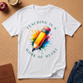 White T-Shirt with text saying "Teaching is a Work of Heart" surrounding a pencil graphic with watercolor overspray 