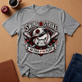 A heather gray short-sleeve t-shirt with a baseball graphic print reading 'SWING OR STRIKE' and 'NO FEAR AT THE PLATE', laid flat on a cork background with a coffee cup, apple, and jeans nearby.
