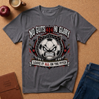 A heather dark gray graphic t-shirt laid flat on a cork background, surrounded by lifestyle props including an apple, a coffee cup, a journal, and jeans. The graphic features a soccer ball with an angry face inside a web, with the text 'NO GUTS 4:24 NO GLORY' above and 'LEAVE IT ALL ON THE PITCH' below.