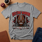 A heather gray graphic t-shirt featuring a cartoon football mascot with the text 'RED ZONE WARRIOR' and 'LEAVE IT ALL ON THE FIELD', styled in a flat-lay on a cork background with a coffee cup, apple, and jeans nearby.