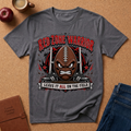 A heather dark gray graphic t-shirt laid flat on a cork background, featuring a football-themed design with the text 'RED ZONE WARRIOR' and 'LEAVE IT ALL ON THE FIELD'. The shirt is styled with a brown leather journal, an apple, a coffee mug, and blue jeans.