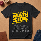 A black graphic t-shirt laid flat on a cork background, featuring a yellow and white design that reads 'COME TO THE MATH SIDE WE HAVE π' with pi digits printed below. Surrounding props include a coffee cup, an apple, a notebook, and jeans.