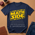 A navy blue graphic t-shirt laid flat on a cork background, featuring a yellow and white design that reads 'COME TO THE MATH SIDE WE HAVE π' with pi digits printed below. Styled flat-lay props include an apple, a coffee cup, a brown journal, and blue jeans.