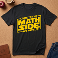 black graphic t-shirt laid flat on a brown cork background, featuring a yellow graphic that reads 'COME TO THE MATH SIDE WE HAVE π' styled with school and coffee props.
