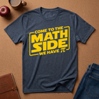 A heather navy blue graphic t-shirt laid flat on a brown cork background, featuring a yellow graphic that reads 'COME TO THE MATH SIDE WE HAVE π' styled with school and coffee props.