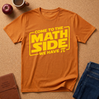 An orange crewneck t-shirt with a yellow graphic reading 'COME TO THE MATH SIDE WE HAVE π' laid flat on a cork background with lifestyle props including an apple, a coffee mug, a notebook, and blue jeans.