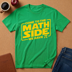 A bright green short-sleeve T-shirt laid flat on a cork surface, printed with large yellow text reading 'COME TO THE MATH SIDE WE HAVE πέε.' Surrounding props include a coffee mug, apple, brown notebook, folded jeans, and a small plant, but no person or real environment is present, so this is Product/Packaging rather than Lifestyle.