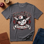 A dark gray graphic t-shirt featuring a baseball design with the text 'Swing or Strike' and 'No Fear at the Plate'. The shirt is displayed in a flat-lay arrangement on a cork background with props including an apple, a coffee mug, a journal, and jeans.