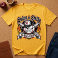 A yellow graphic t-shirt laid flat on a cork background, featuring a baseball-themed 'Swing or Strike No Fear at the Plate' design. The shirt is styled with lifestyle props including an apple, a coffee mug, a brown leather notebook, and blue jeans.