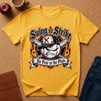 A yellow graphic t-shirt laid flat on a cork background, featuring a baseball-themed 'Swing or Strike No Fear at the Plate' design. The shirt is styled with lifestyle props including an apple, a coffee mug, a brown leather notebook, and blue jeans.