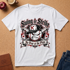 A white short-sleeve graphic T-shirt laid flat on a cork background, featuring a baseball-themed distressed print with the words 'Swing or Strike' and 'No Fear at the Plate.' The shirt is styled with a red apple, a mug, a journal, and jeans around it.