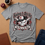 A heather gray graphic t-shirt laid flat on a brown cork background, featuring a baseball-themed 'Swing or Strike No Fear at the Plate' design. The shirt is styled with a coffee cup, an apple, a notebook, and jeans.