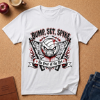 A white short-sleeve graphic T-shirt laid flat on a cork background, featuring a volleyball-themed angry face design with the text 'BUMP, SET, SPIKE' and 'No Fear of the Net.' Styled flat-lay props include a red apple, a coffee cup, a brown journal, and blue jeans.