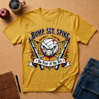 A yellow graphic t-shirt laid flat on a cork background, surrounded by lifestyle props including a coffee mug, an apple, a notebook, a pen, and folded blue jeans. The t-shirt features a volleyball-themed graphic with the text 'BUMP, SET, SPIKE' and 'No Fear of the Net'.