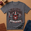 A dark gray graphic t-shirt laid flat on a cork background, featuring a football helmet with a red snarling face and the text 'RED ZONE WARRIOR' and 'Leave it All on the Field'. Props like an apple, a mug, a journal, and jeans are visible around it.