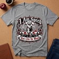 A heather gray short-sleeve t-shirt with a detailed graphic print of a cheetah running with the text 'I Am Speed' and 'Run Like the Wind', displayed flat on a cork background with lifestyle props like an apple, coffee cup, notebook, and jeans.