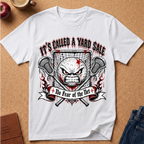 A white short-sleeve graphic T-shirt laid flat on a cork surface, featuring a large distressed design with the text 'IT'S CALLED A YARD SALE' and 'No Fear of the Net' around a stylized angry face, lacrosse sticks, and a goal net. Partial props including an apple, a cup, a book, and jeans are visible around the shirt.