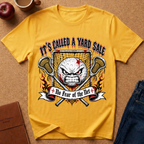 A yellow graphic t-shirt laid flat on a brown cork-like surface, surrounded by lifestyle props including a red apple, a coffee mug, a brown journal, and blue jeans. The t-shirt features a graphic of an angry lacrosse ball behind crossed lacrosse sticks and a net, with the text 'IT'S CALLED A YARD SALE' and a banner reading '+ No Fear of the Net +'.