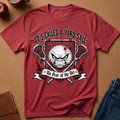 A heathered red graphic t-shirt laid flat on a brown cork background, surrounded by lifestyle props like jeans, a notebook, and a apple. The shirt features a graphic of an angry soccer ball with a star, crossed lacrosse sticks, and the text 'IT'S CALLED A YARD SALE' and '+ No Fear of the Net +'.
