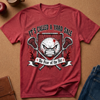 A heathered red graphic t-shirt laid flat on a brown cork background, surrounded by lifestyle props like jeans, a notebook, and a apple. The shirt features a graphic of an angry soccer ball with a star, crossed lacrosse sticks, and the text 'IT'S CALLED A YARD SALE' and '+ No Fear of the Net +'.