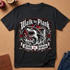 A black graphic t-shirt laid flat on a cork background, featuring a large shark design with the text 'Walk the Plank Sink or Swim'.