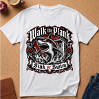 A white short-sleeve graphic T-shirt laid flat on a cork background, featuring a large shark illustration and the text 'Walk the Plank Sink or Swim' in black, red, and white.