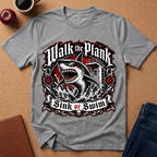 A heather gray short-sleeve t-shirt with a large graphic print of a shark and the words 'Walk the Plank Sink or Swim', laid flat on a cork background with props including an apple, coffee cup, notebook, and jeans.