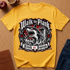 A yellow short-sleeve graphic T-shirt laid flat on a cork-like surface, featuring a large shark illustration and the text 'Walk the Plank Sink or Swim.' Surrounding props include a cup, apple, notebook, and jeans, but no person or real environment is present, so this is Product/Packaging rather than Lifestyle.