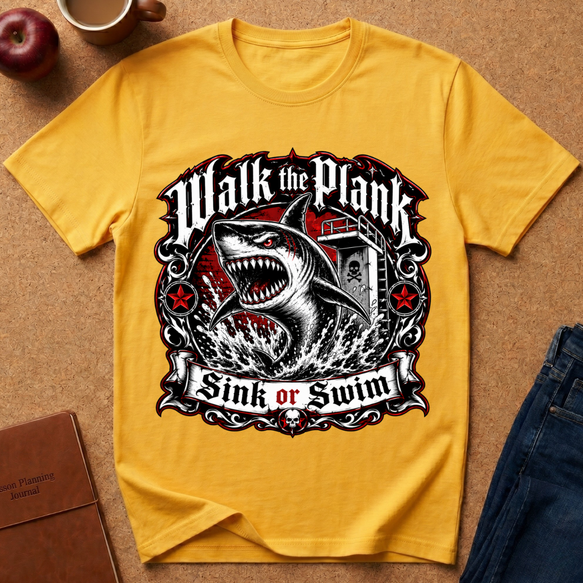 A yellow short-sleeve graphic T-shirt laid flat on a cork-like surface, featuring a large shark illustration and the text 'Walk the Plank Sink or Swim.' Surrounding props include a cup, apple, notebook, and jeans, but no person or real environment is present, so this is Product/Packaging rather than Lifestyle.