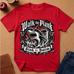 A red short-sleeve graphic T-shirt laid flat on a cork-like surface, featuring a large shark illustration and the words 'Walk the Plank Sink or Swim' in a black-and-white ornate emblem. Partial props around the shirt include an apple, a mug, a brown notebook, and blue jeans.