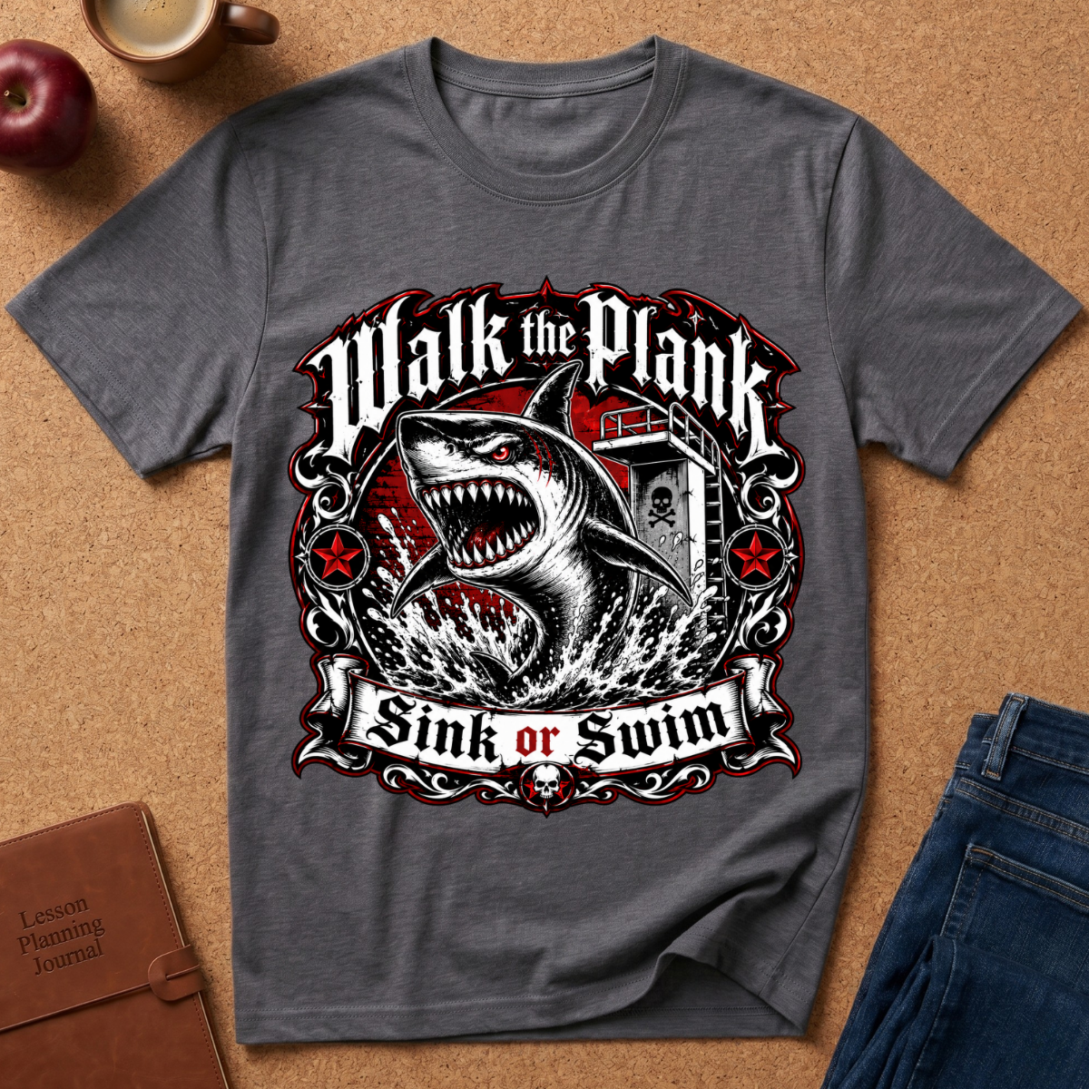 A dark gray graphic t-shirt laid flat on a cork background, featuring a shark and anchor design with the text 'Walk the Plank Sink or Swim'. The shirt is styled with props including an apple, a coffee cup, a journal, and blue jeans.