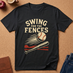 A black short-sleeve graphic T-shirt laid flat on a cork background, featuring the phrase 'SWING FOR THE FENCES' above a baseball and bat illustration. The shirt is styled with partial props including an apple, a cup, a journal, and jeans.