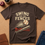 A dark brown graphic t-shirt laid flat on a cork background, featuring a baseball design with the text 'SWING FOR THE FENCES'. The shirt is styled with a coffee mug, an apple, a notebook, and blue jeans.