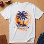 A white short-sleeve crewneck t-shirt laid flat on a cork background, featuring a graphic print of a palm tree, a chair, and the words 'Summer Season' in orange and navy blue.