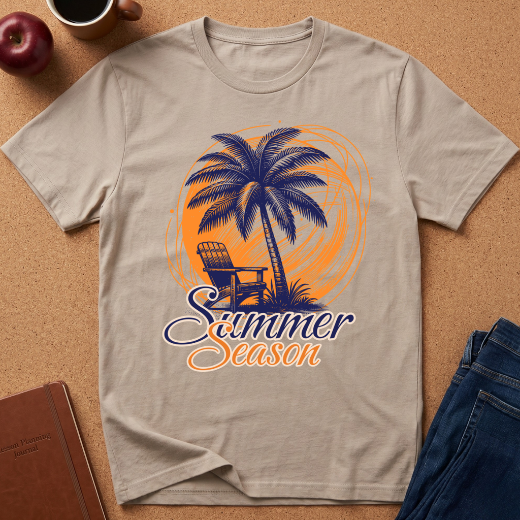 A beige graphic t-shirt laid flat on a cork background, featuring a blue palm tree, chair, and orange circular design with the text 'Summer Season'. Props like an apple, coffee cup, book, and jeans are visible around it.