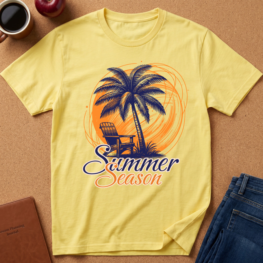 A pale yellow graphic T-shirt laid flat on a cork surface, featuring a palm tree, chair, orange circular design, and the words 'Summer Season.' Surrounding props include a coffee cup, apple, notebook, and folded jeans.