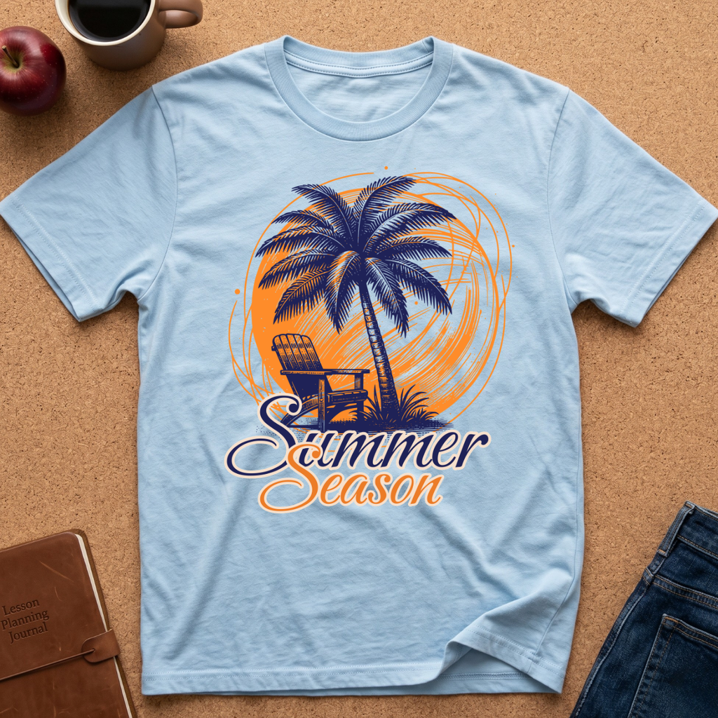 A light blue short-sleeve t-shirt laid flat on a cork surface, featuring a graphic of a palm tree, beach chair, and orange circular design with the words 'Summer Season.' Partial props around it include a mug, apple, notebook, and jeans.