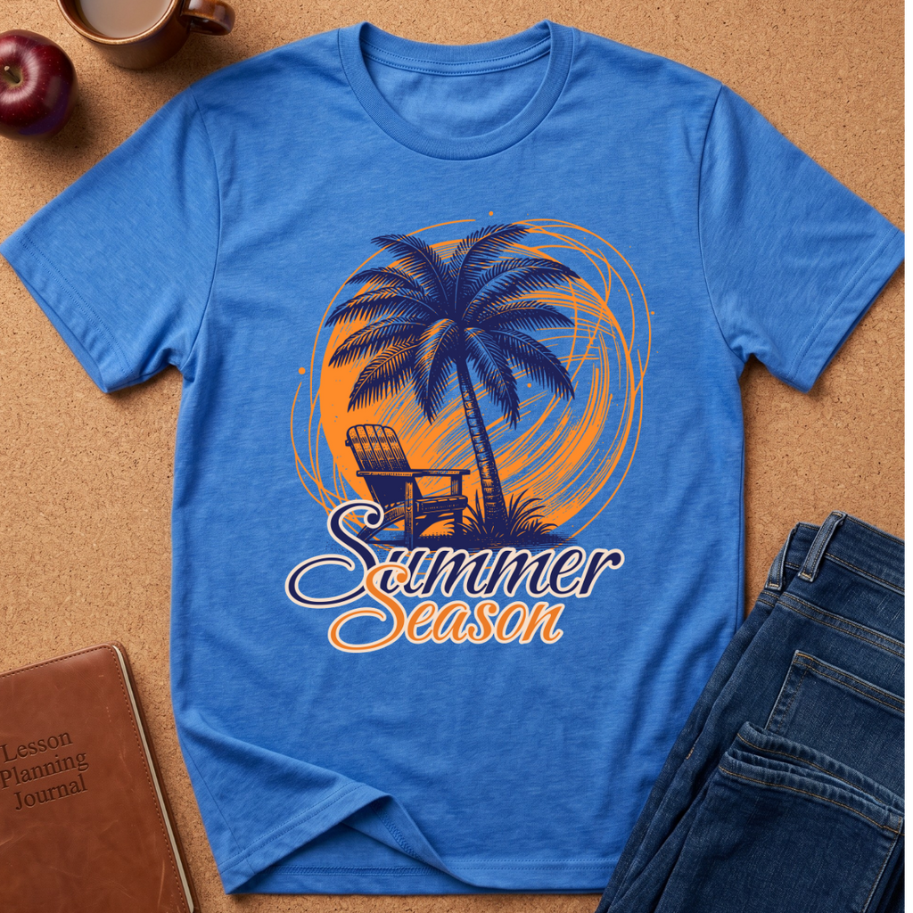 A blue short-sleeve graphic T-shirt laid flat on a cork-like surface, featuring a palm tree, chair, and the words 'Summer Season' in orange and white. Surrounding props include a brown journal, a cup, an apple, and folded blue jeans.