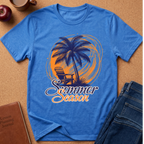 A blue short-sleeve graphic T-shirt laid flat on a cork-like surface, featuring a palm tree, chair, and the words 'Summer Season' in orange and white. Surrounding props include a brown journal, a cup, an apple, and folded blue jeans.