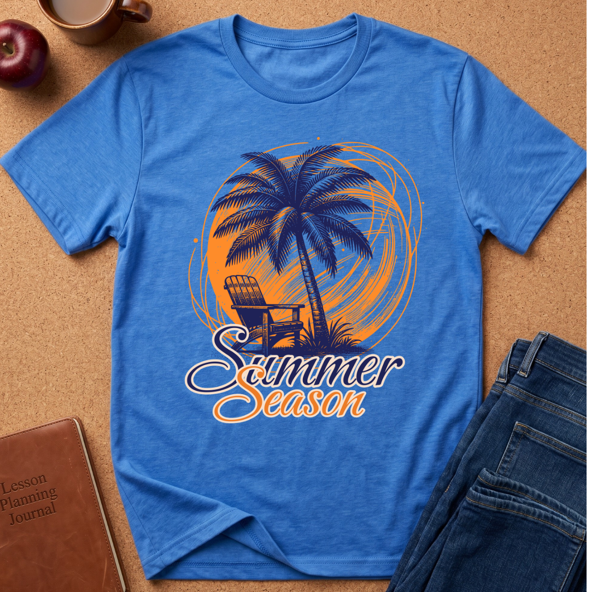 A blue short-sleeve graphic T-shirt laid flat on a cork-like surface, featuring a palm tree, chair, and the words 'Summer Season' in orange and white. Surrounding props include a brown journal, a cup, an apple, and folded blue jeans.