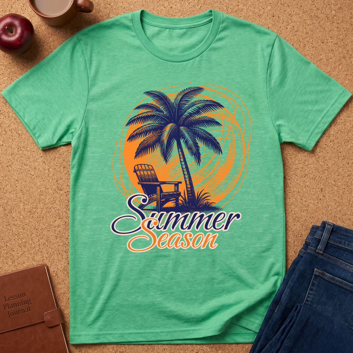 A green short-sleeve graphic T-shirt laid flat on a cork surface, featuring a palm tree, chair, and the words 'Summer Season' in orange and purple. Surrounding props include an apple, coffee cup, brown journal, and blue jeans.