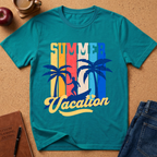 A teal short-sleeve graphic T-shirt laid flat on a cork-like surface, surrounded by lifestyle props including an apple, coffee cup, notebook, and jeans. The shirt features a retro vertical stripe design in yellow, orange, red, and blue with palm trees and the words 'SUMMER Vacation' in yellow script.